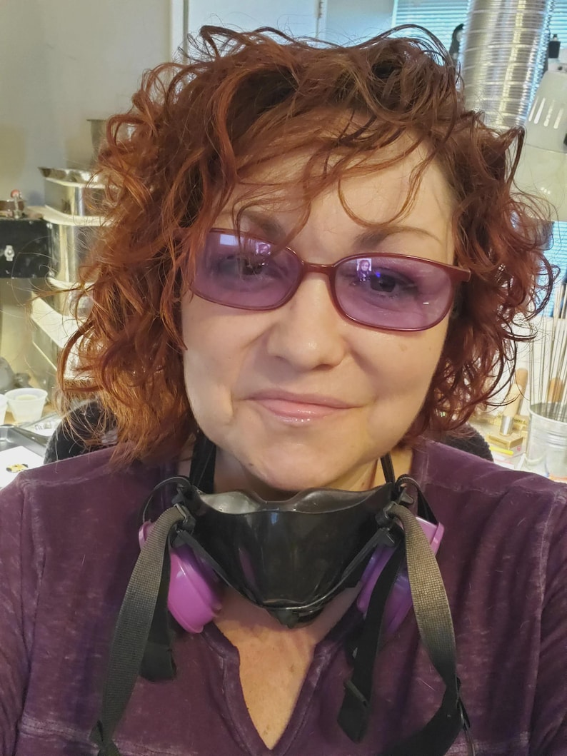 May include: A person with curly auburn hair wearing purple-tinted glasses and a black respirator mask with purple filter cartridges. The individual is dressed in a purple long-sleeved shirt, with the mask resting around the neck area.