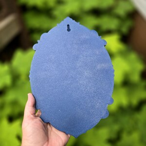 May include: A blue, oval-shaped decorative plaque with a textured, matte finish and ornate, scalloped edges. The object features a single, centered keyhole-style mounting slot at the top, designed for hanging on a wall surface.