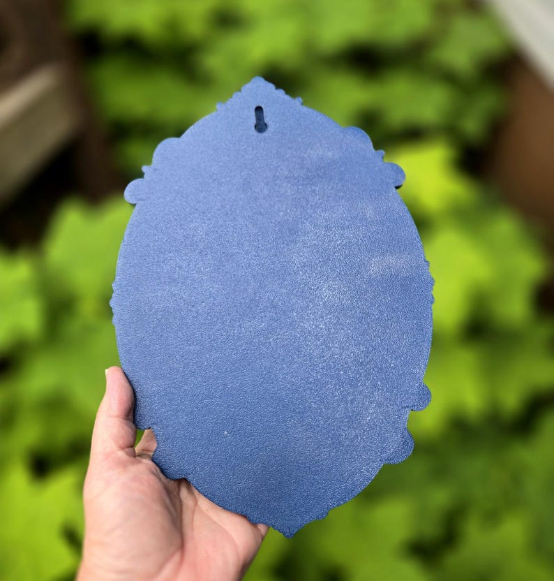 May include: A blue, oval-shaped decorative plaque with a textured, matte finish and ornate, scalloped edges. The object features a single, centered keyhole-style mounting slot at the top, designed for hanging on a wall surface.