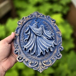 May include: An oval wall plaque featuring a relief of two figures draped in flowing fabric, set against a background of ferns and a crescent moon. The piece is finished in a deep blue with gold highlights on the ornate, scrolled frame edges.