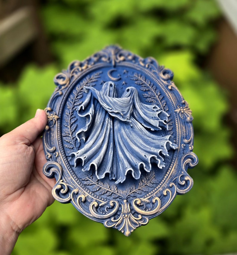 May include: An oval wall plaque featuring a relief of two figures draped in flowing fabric, set against a background of ferns and a crescent moon. The piece is finished in a deep blue with gold highlights on the ornate, scrolled frame edges.