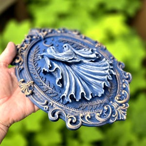 May include: An oval wall plaque featuring a relief sculpture of two ghosts with flowing robes. The piece is finished in a deep blue with gold-toned highlights on the ornate, scrollwork frame and the edges of the ghostly figures' garments.