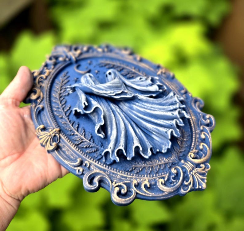 May include: An oval wall plaque featuring a relief sculpture of two ghosts with flowing robes. The piece is finished in a deep blue with gold-toned highlights on the ornate, scrollwork frame and the edges of the ghostly figures' garments.