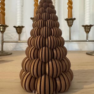 May include: A brown, layered, wooden Christmas tree-shaped decoration. The tree is made of stacked, rounded, horizontal pieces. Behind the tree are several candles in brass candle holders. The candles are a mix of spiral and straight designs.