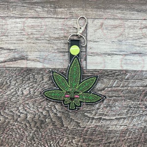 Puede incluir: Un llavero de hoja de cannabis verde con borde negro y costuras blancas. La hoja tiene una cara con ojos rosas y una boca sonriente. El llavero tiene un cierre plateado y una correa negra con un botón verde neón.