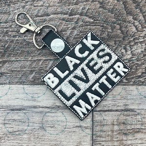 May include: Black Lives Matter keychain with a silver-tone clasp. The black and white embroidered design features the phrase "BLACK LIVES MATTER" in bold, capital letters. The keychain includes a small black strap with the text "TCH'D MLXVR".