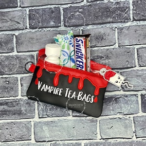 May include: A black and red zippered pouch with the text "VAMPIRE TEA BAGS". The pouch contains tea bags, a bottle, and a Snickers bar. A small tag with a cartoon character is attached to the zipper.