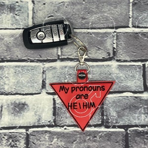May include: A red, triangular keychain with the text "My pronouns are HE/HIM" in white. The keychain is attached to a silver clasp and a car key. The background is a gray brick wall.
