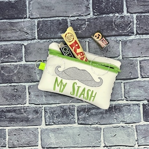 Puede incluir: Bolsa blanca con cremallera verde, con diseño de bigote y texto bordado "MY STASH" en verde. Contiene un encendedor, papel de liar y filtros. Se adjunta un pequeño llavero.