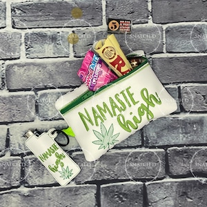 Könnte beinhalten: Weiße Reißverschlusstasche mit grünen Verzierungen und den Worten "NAMASTE high" in Grün, zusammen mit einem Cannabisblatt-Design. Enthält ein passendes Feuerzeug und Zubehör.