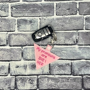 May include: A pink, triangular keychain with the text "My pronouns are SHE / HER" in a white stitched font. The keychain is attached to a car key fob with a silver keyring.