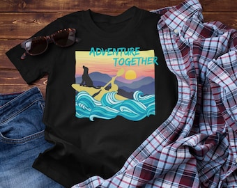 Puede incluir: Camiseta negra con un gráfico colorido de una persona en kayak con un perro, el texto "ADVENTURE TOGETHER" está encima de la imagen. La camiseta se muestra con gafas de sol, vaqueros y una camisa a cuadros.
