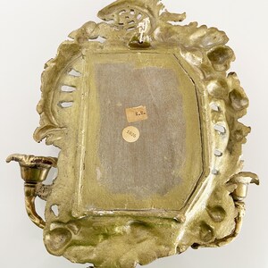 May include: A gold-toned metal wall sconce with ornate detailing and two candle holders. The back of the sconce has a label with the text "K.B. 1826".