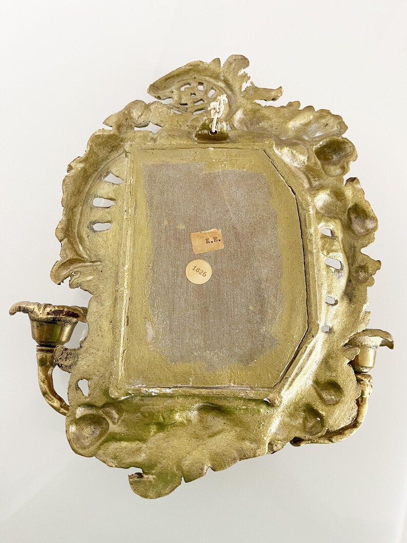May include: A gold-toned metal wall sconce with ornate detailing and two candle holders. The back of the sconce has a label with the text "K.B. 1826".