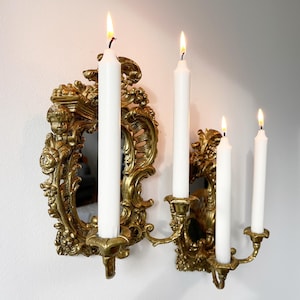 May include: Gold wall-mounted candle holder with ornate details and a mirror. Four white candles are lit.