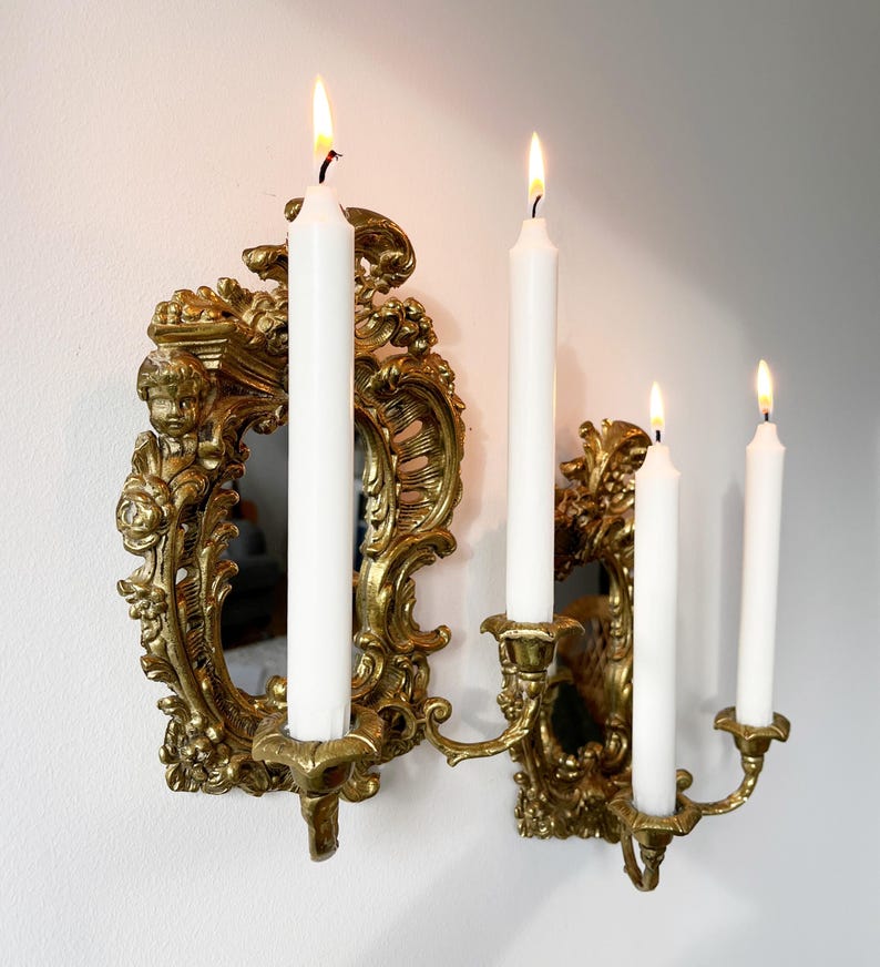 May include: Gold wall-mounted candle holder with ornate details and a mirror. Four white candles are lit.