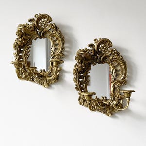 May include: A pair of ornate gold-toned wall sconces with mirrored centers. Each sconce features intricate scrollwork and floral designs.
