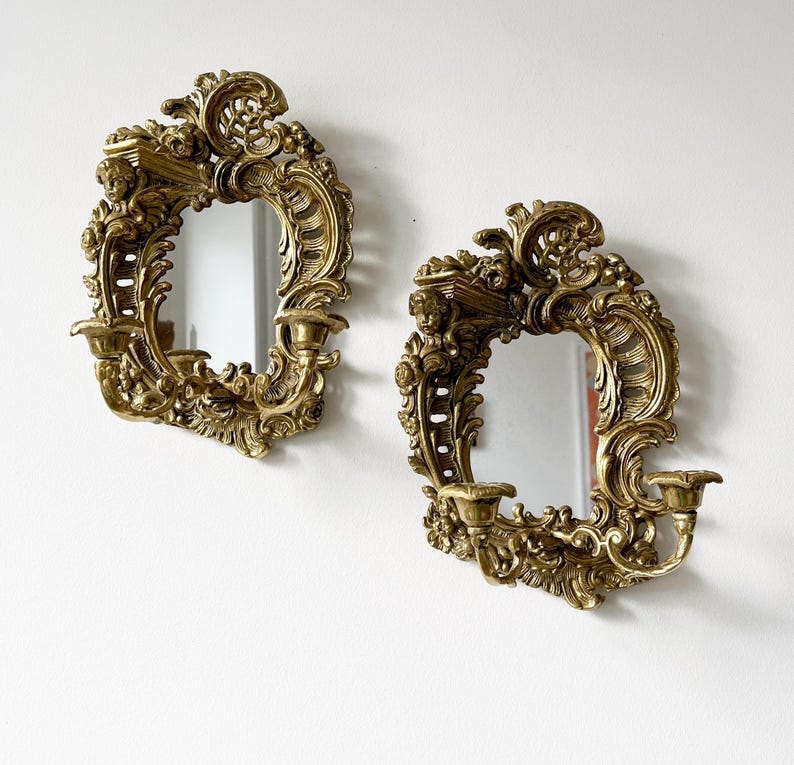 May include: A pair of ornate gold-toned wall sconces with mirrored centers. Each sconce features intricate scrollwork and floral designs.