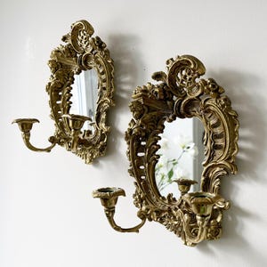 May include: A pair of ornate gold-toned wall sconces with mirrors and candle holders. Each sconce features intricate scrollwork and a beveled mirror.