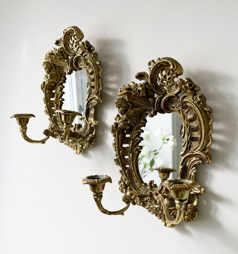May include: A pair of ornate gold-toned wall sconces with mirrors and candle holders. Each sconce features intricate scrollwork and a beveled mirror.