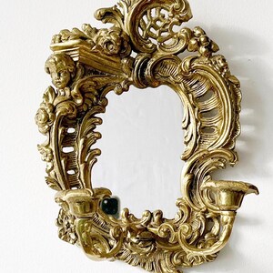 May include: A gold-toned ornate wall mirror with a decorative frame featuring a cherub and scrollwork. The mirror has a candle holder on each side.