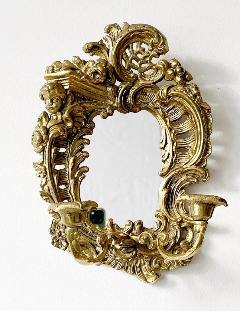 May include: A gold-toned ornate wall mirror with a decorative frame featuring a cherub and scrollwork. The mirror has a candle holder on each side.