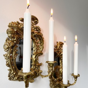 May include: A gold ornate wall-mounted candle holder with a mirror in the center. Four white candles are lit in the candle holders.