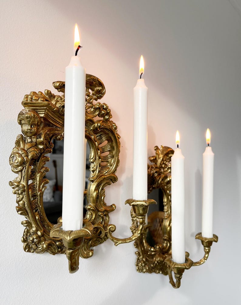 May include: A gold ornate wall-mounted candle holder with a mirror in the center. Four white candles are lit in the candle holders.