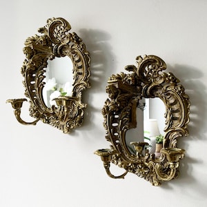 May include: A pair of ornate gold-toned wall sconces with mirrored centers and candle holders. The sconces feature intricate floral and scrollwork designs.
