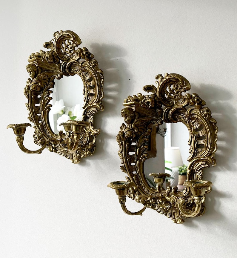 May include: A pair of ornate gold-toned wall sconces with mirrored centers and candle holders. The sconces feature intricate floral and scrollwork designs.