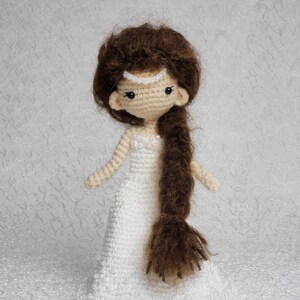 May include: A crocheted doll wearing a white beaded dress and a pearl headband. The doll has brown hair styled in a long braid.