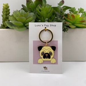 May include: A gold key chain with a cartoon pug face charm. The charm is on a pink background with the text "Luna's Pug Shop" and "A Pug Helping Pugs".