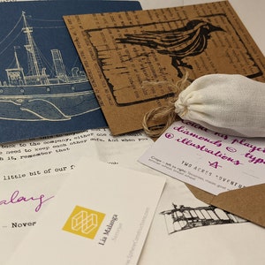 Puede incluir: Un surtido de artículos de papelería, que incluyen una tarjeta azul marino con una ilustración de un barco, una tarjeta marrón con un pájaro y una pequeña bolsa con cordón. También hay varias notas manuscritas y un sobre marrón.