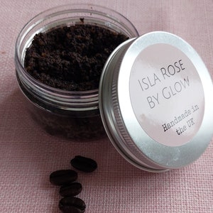 Puede incluir: Un tarro de vidrio transparente con tapa plateada que contiene un exfoliante de café marrón oscuro. La tapa tiene una etiqueta blanca con el texto "Isla Rose by Glow" y "Handmade in the UK". Hay cuatro granos de café sobre una superficie rosa.