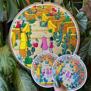 May include: An embroidered artwork in a wooden hoop depicts two figures walking through a maze-like structure. The artwork features yellow, pink, and green colors. Two matching stickers are also visible, held by a hand with green nail polish.