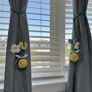 Könnte beinhalten: Zwei graue Vorhänge, die mit grünen Seilen zurückgebunden sind und jeweils mit gehäkelten Bienen und Blumen verziert sind. Einer hat eine Biene mit einer weißen Gänseblümchen und gelben Blume. Der andere hat eine Biene mit einem blauen Schmetterling und einer weißen Blume.