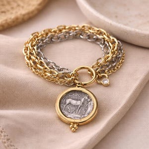 May include: A gold and silver layered bracelet with a round pendant. The pendant features a silver coin design with a horse image. The bracelet has a gold clasp and a small, clear charm. The jewellery is displayed on a beige fabric.