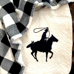 May include: A white and black buffalo plaid blanket with a white sherpa backing. The sherpa side features a black silhouette of a cowboy riding a horse.
