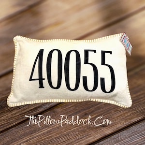 May include: A white pillow with a black number "40055" printed on it. The pillow has a gold stitched border and is sitting on a wooden surface.