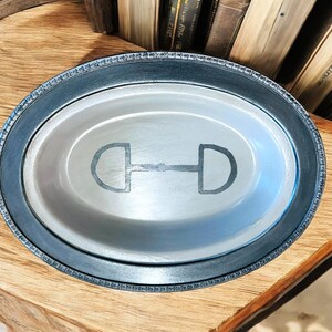 Könnte beinhalten: Eine ovale Servierplatte aus Silber mit schwarzem Rand und einem silbernen Steigbügel-Design in der Mitte.