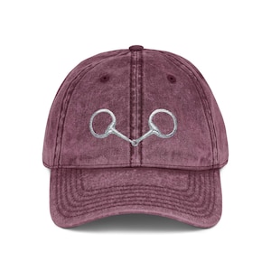 May include: A maroon baseball cap with a silver-colored horse bit embroidered on the front. The cap has a curved brim and a vintage, worn look.