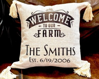 Könnte beinhalten: Ein beiges quadratisches Kissen mit dem Text "WELCOME TO OUR FARM The Smiths Est. 6/19/2006" in Schwarz. Das Kissen hat vier cremefarbene Quasten, eine an jeder Ecke. Das Kissen liegt auf einer braunen Lederoberfläche.