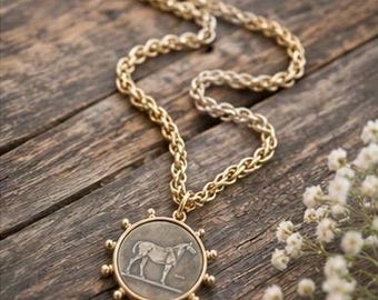 Può includere: Collana color oro con un ciondolo circolare raffigurante un cavallo. Il ciondolo è circondato da piccole borchie decorative. La catena ha un design spesso e strutturato. La collana è appoggiata su una superficie di legno.