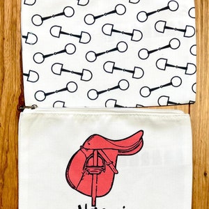 Puede incluir: Dos bolsas de cremallera de lona blanca. La bolsa superior tiene un patrón repetido en blanco y negro de bocados de caballo. La bolsa inferior tiene una silla de montar roja y el nombre Naomi impreso en negro.