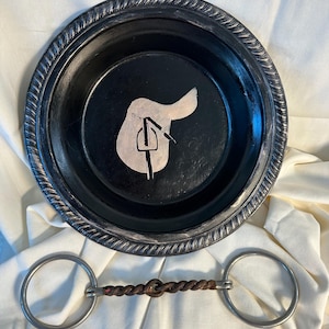 May include: A black and silver decorative plate with a white silhouette of a saddle and stirrups. A silver and brown twisted bit with two silver rings is also pictured.