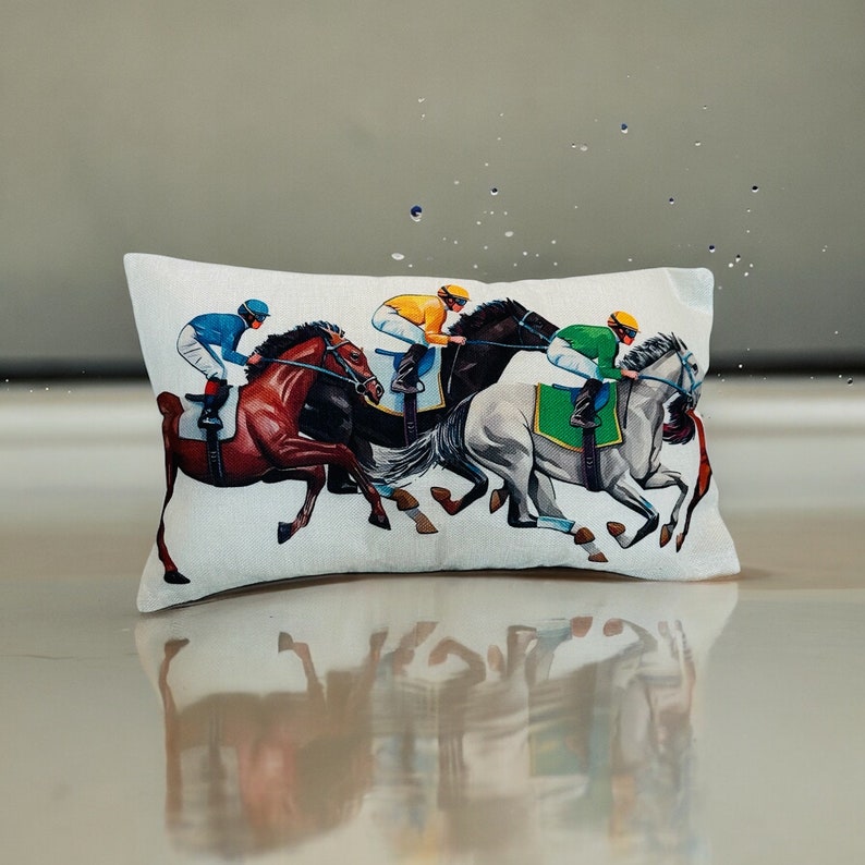May include: A white pillow with a colorful illustration of four horses and riders. The horses are brown, black, and white, and the riders are wearing colorful jockey outfits. The pillow is a decorative accent for a home.