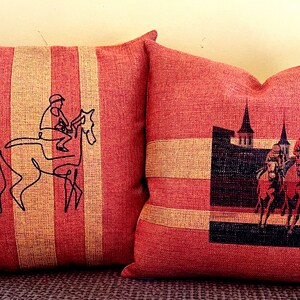 May include: Two decorative throw pillows with a red and beige striped pattern. One pillow features a black line drawing of a jockey on a horse. The other pillow has a graphic of jockeys on horses and a building.