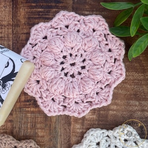 May include: A pink crocheted coaster with a delicate floral design. The coaster is made with a soft yarn and has a textured surface.