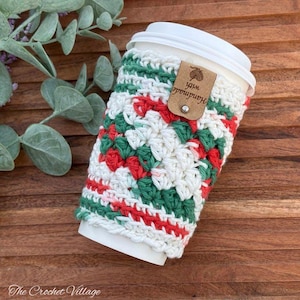 May include: A white disposable coffee cup featuring a crocheted sleeve with a red, green, and white granny square pattern. The sleeve includes a tan leather-like tag with the text "Handmade with" and a small yarn ball icon, secured by a silver stud.