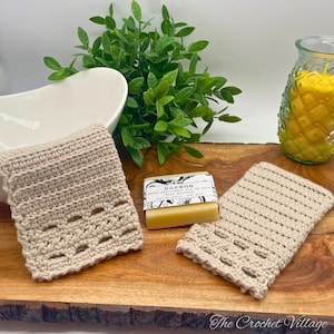 May include: Two crocheted soap savers in beige, one with a more intricate design, the other with a simple ribbed pattern. A bar of soap with the text "Sorene" is on top of the ribbed soap saver. The soap savers are on a wooden surface.